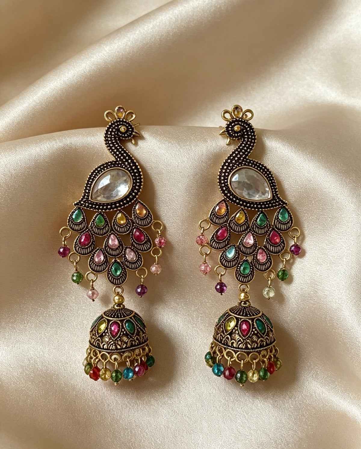 Flat lay view of Majestic Peacock Heritage antique gold-plated multistone jhumka earrings displayed on a satin fabric background, highlighting the intricate peacock design and colorful beaded fringes.