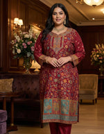 A model standing in a luxury interior wearing a Deep Maroon Pure Silk kurta set with intricate multi-colored Kanni embroidery, zari work, and pure raw silk trousers.