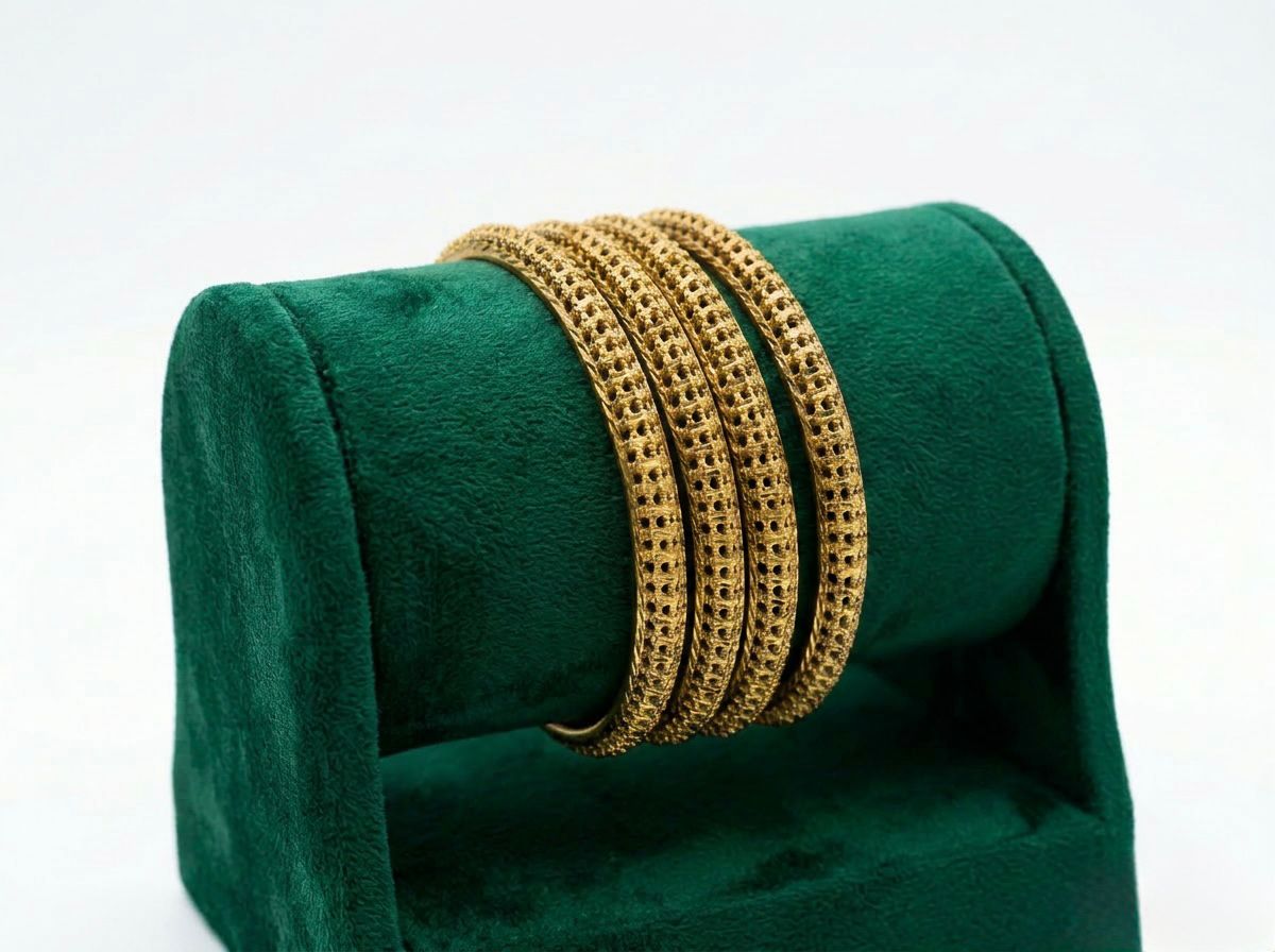 A professional product photograph of four micro gold-plated copper bangles displayed on a green velvet jewelry roll. The bangles feature an intricate, repetitive honeycomb-style textured pattern with a high-shine finish, set against a clean white background.