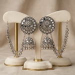 A pair of ornate silver-toned Indian jhumka earrings displayed on cream-colored velvet jewelry stands with gold-colored metal supports against a soft beige background.