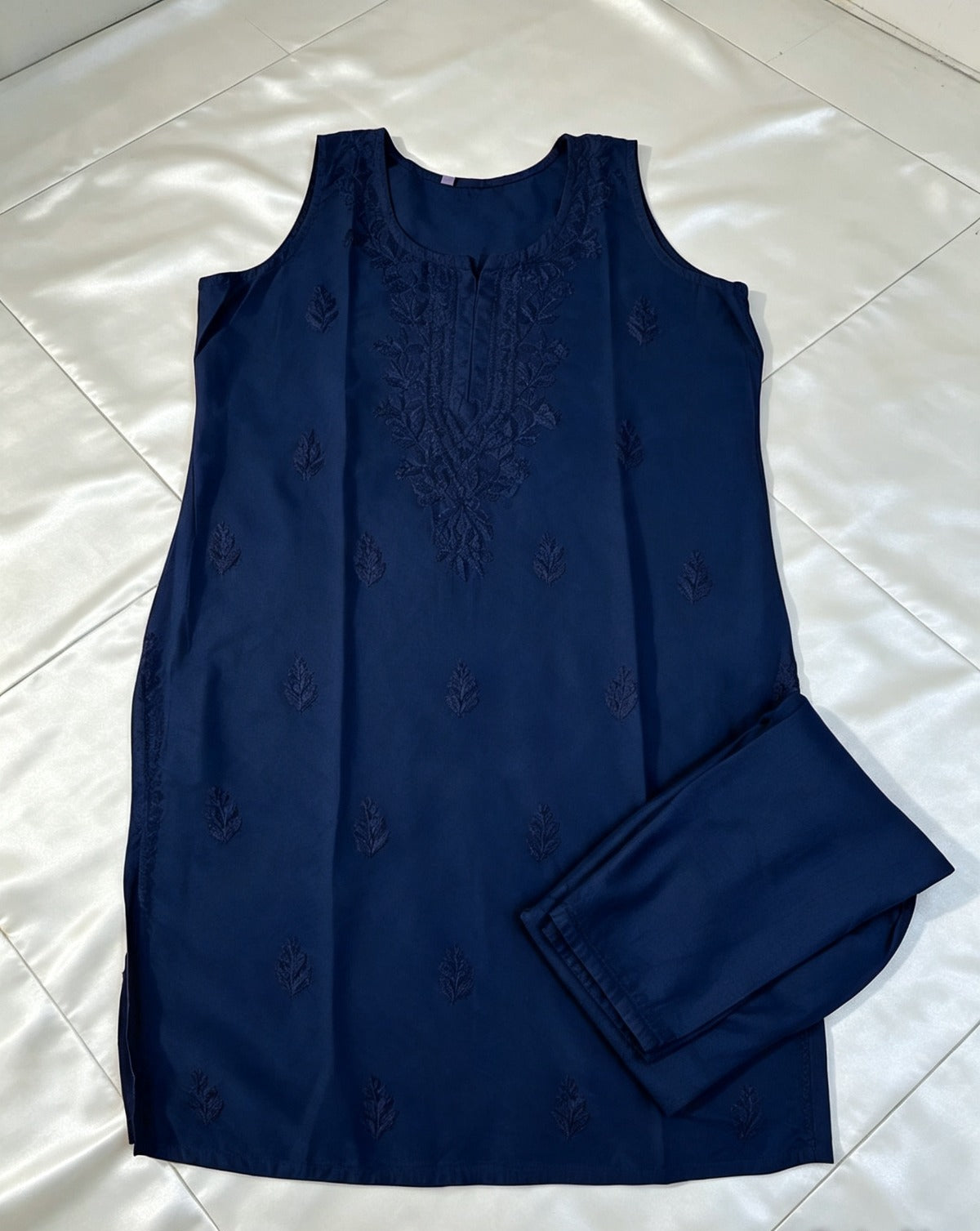 Flat lay view of the navy blue handcrafted sleeveless Chanderi silk Chikankari tunic and matching folded trousers neatly arranged on a glossy white background.