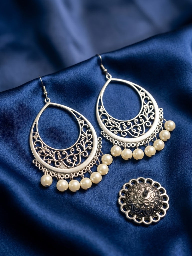 Silver-toned crescent-shaped filigree earrings with dangling cream pearl beads, displayed on a dark blue satin fabric alongside a matching round earring back.