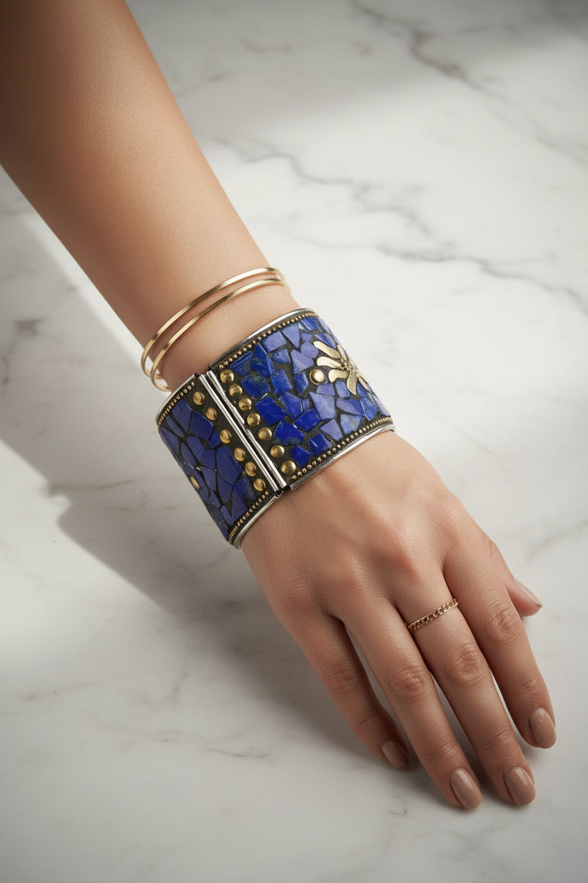 A close-up of a person's hand and wrist wearing a wide blue mosaic cuff bracelet with gold-toned metal details, alongside two slim gold bangles, set against a dark brown textured background with scattered lapis lazuli stones.