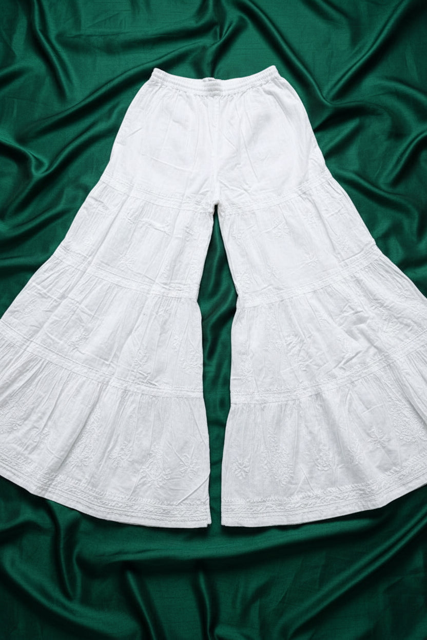 Flat-lay view of a white cotton 4-tier flared sharara featuring intricate Chikankari embroidery, laid out on a dark background.