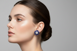 A close-up side profile of a woman with dark hair in a low bun, wearing a blue and white flower-shaped stud earring against a solid gray background.