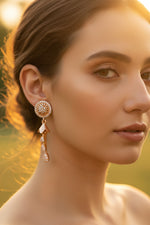 Model wearing golden brass earrings featuring a circular floral stud encrusted with crystals and a three-tier crystal teardrop dangle.