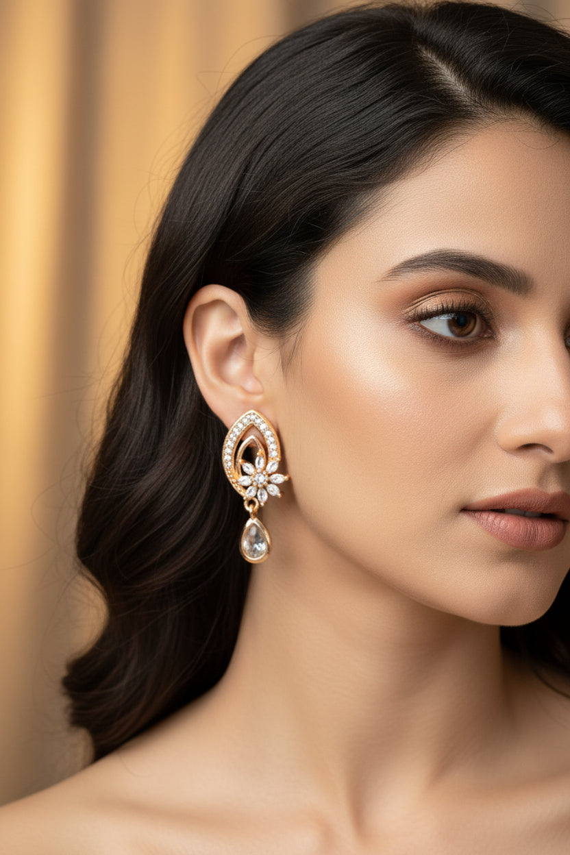 Model wearing rose gold brass earrings featuring an intricate, petal-shaped frame encrusted with small crystals and a dangling, clear teardrop charm.