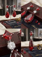 A four-panel collage showing red teardrop and square gemstone drop earrings displayed on a stand, on a dark wooden box, and in close-up detail. The earrings have silver-toned metal settings with clear round stones.