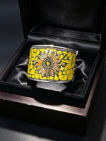Mosaic openable sunflower bracelet. A wide cuff bracelet displayed in an open black presentation box. The bracelet has a mosaic yellow pattern with a central sunflower design in yellow, brown, and black, bordered by silver-toned metal.
