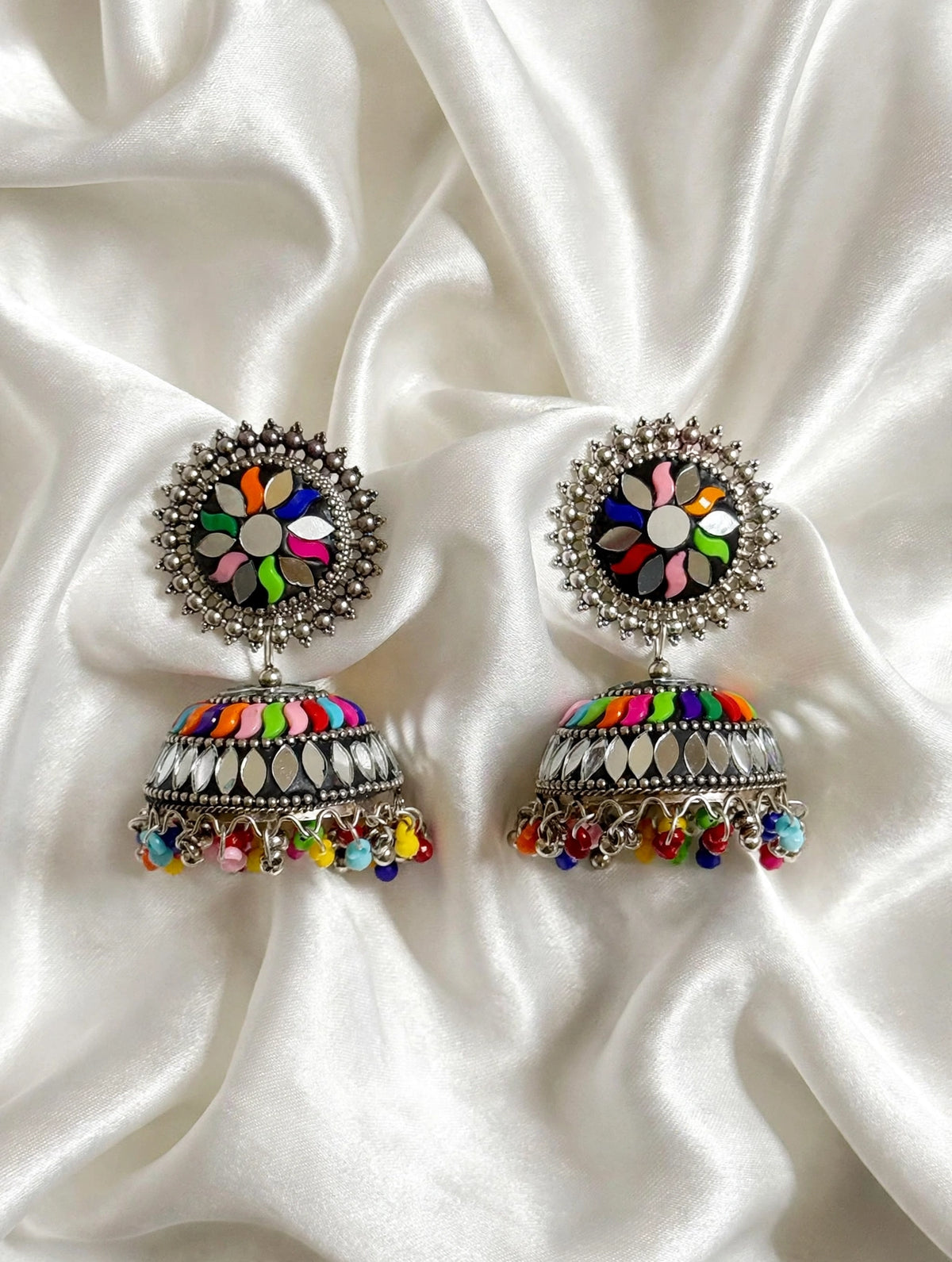 Multicolor floral mirror work meenakari oxidized silver jhumka earrings laid on white silk — Indian Pakistani ethnic bridal party statement jewelry.