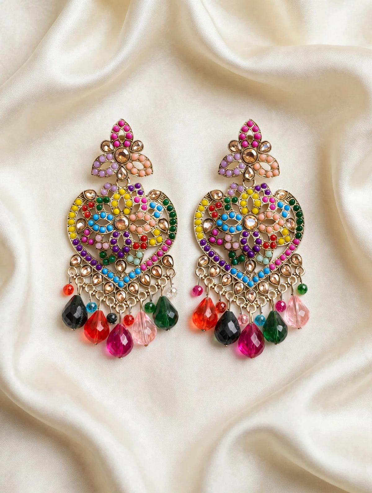 Gold-tone multicolor mosaic heart chandelier earrings with rainbow stones and teardrop bead drops, laid flat on soft cream silk — South Asian bridal and festive jewelry.