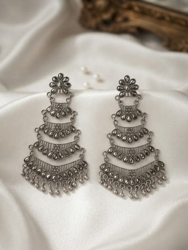 A pair of ornate silver-toned tiered chandelier earrings with dangling bead details, displayed on draped white satin fabric.