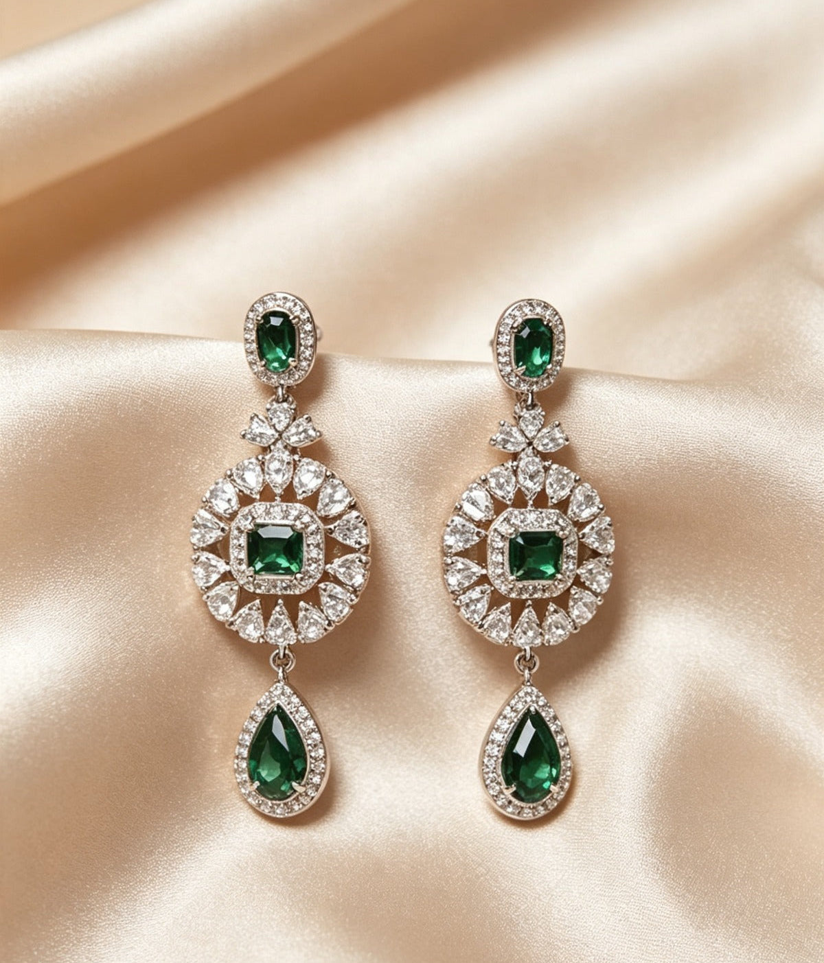 A pair of ornate, silver-tone chandelier earrings featuring a small emerald-cut green stone at the top, a larger central round cluster of clear and emerald-cut green stones, and a large pear-shaped green drop at the bottom, all set against a background of draped champagne-colored satin. The intricate design is symmetrical and catches the light, suggesting a high-quality, diamond-like sparkle.