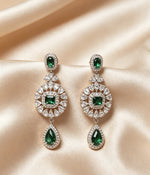 A pair of ornate, silver-tone chandelier earrings featuring a small emerald-cut green stone at the top, a larger central round cluster of clear and emerald-cut green stones, and a large pear-shaped green drop at the bottom, all set against a background of draped champagne-colored satin. The intricate design is symmetrical and catches the light, suggesting a high-quality, diamond-like sparkle.