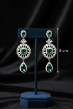 A pair of silver-toned drop earrings with green gemstones and clear stones, displayed on a dark blue stand against a black satin background, with a 5 cm height marker shown on the right.