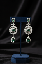 A pair of ornate drop earrings displayed on a blue velvet jewelry stand against a dark draped fabric background. The earrings feature round and teardrop-shaped emerald-cut green gemstones surrounded by small clear stones in a silver-toned setting.