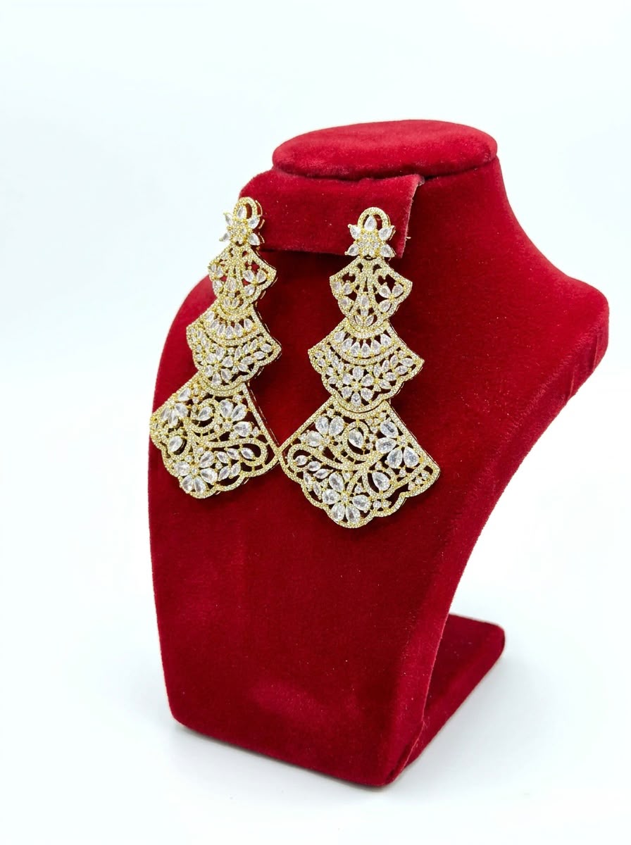 Side view of elegant three-tiered gold-tone statement earrings with detailed filigree patterns and crystal embellishments, shown on a red velvet display stand.