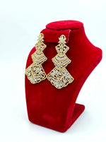 Side view of elegant three-tiered gold-tone statement earrings with detailed filigree patterns and crystal embellishments, shown on a red velvet display stand.