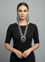 Woman wearing a black outfit with a large turquoise necklace and earrings on a grey background