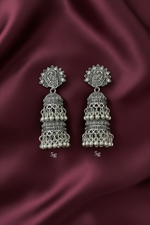 A posh model wearing a pair of intricate, oxidized silver jhumka earrings against a clean, pure white background.