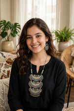 Woman wearing oxidized silver three-tiered multicolour stone statement necklace and jhumka earrings, ethnic Afghan-inspired jewelry with red, green and blue accents, styled with black outfit.