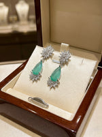 Pair of earrings with green gemstones and diamond-like elements on a display box.