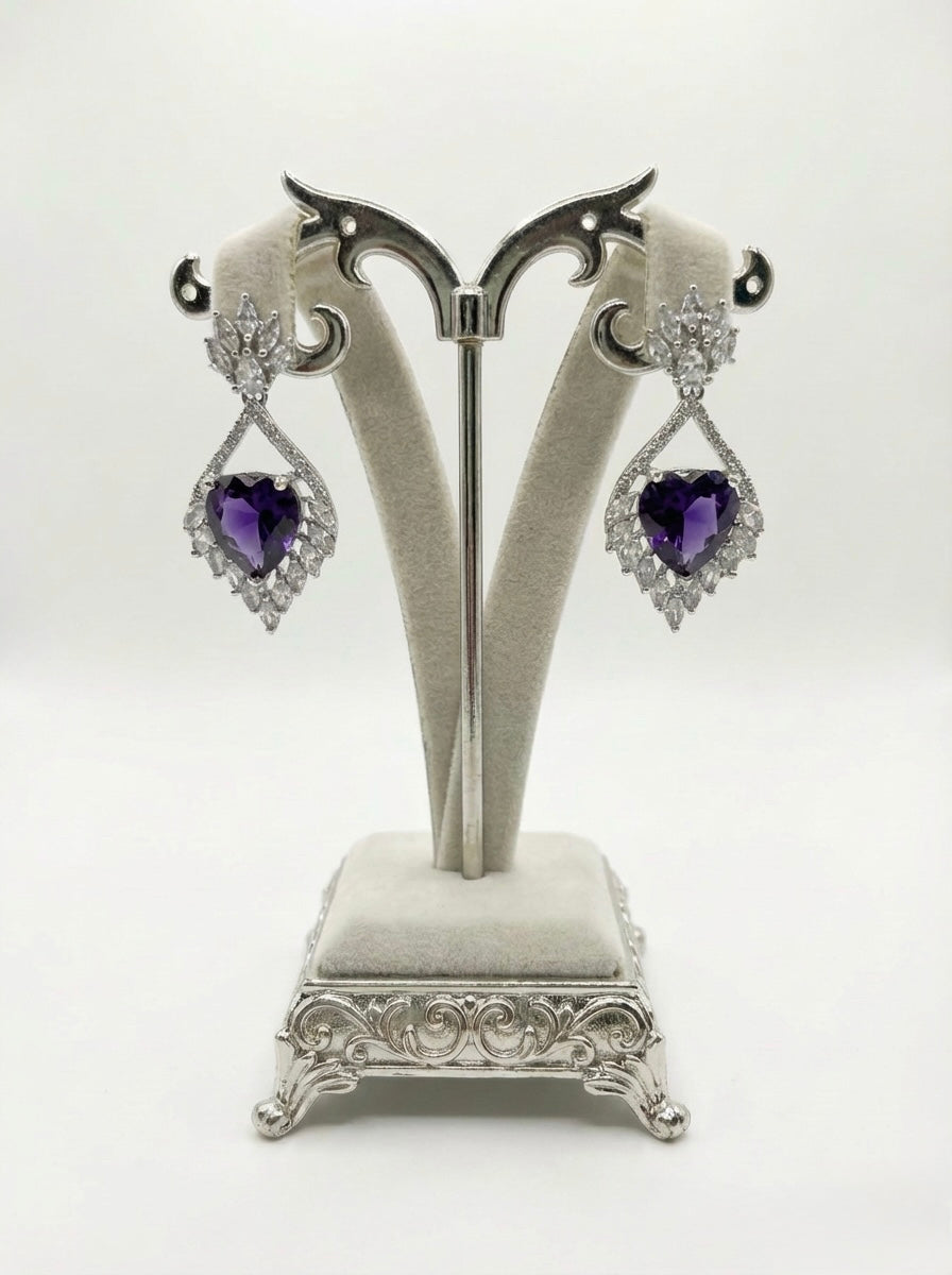 Pair of heart-shaped purple gemstone earrings on a decorative stand against a white background