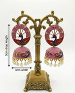 Pair of pink jhumka earrings with peacock design on a decorative stand, showing measurements.