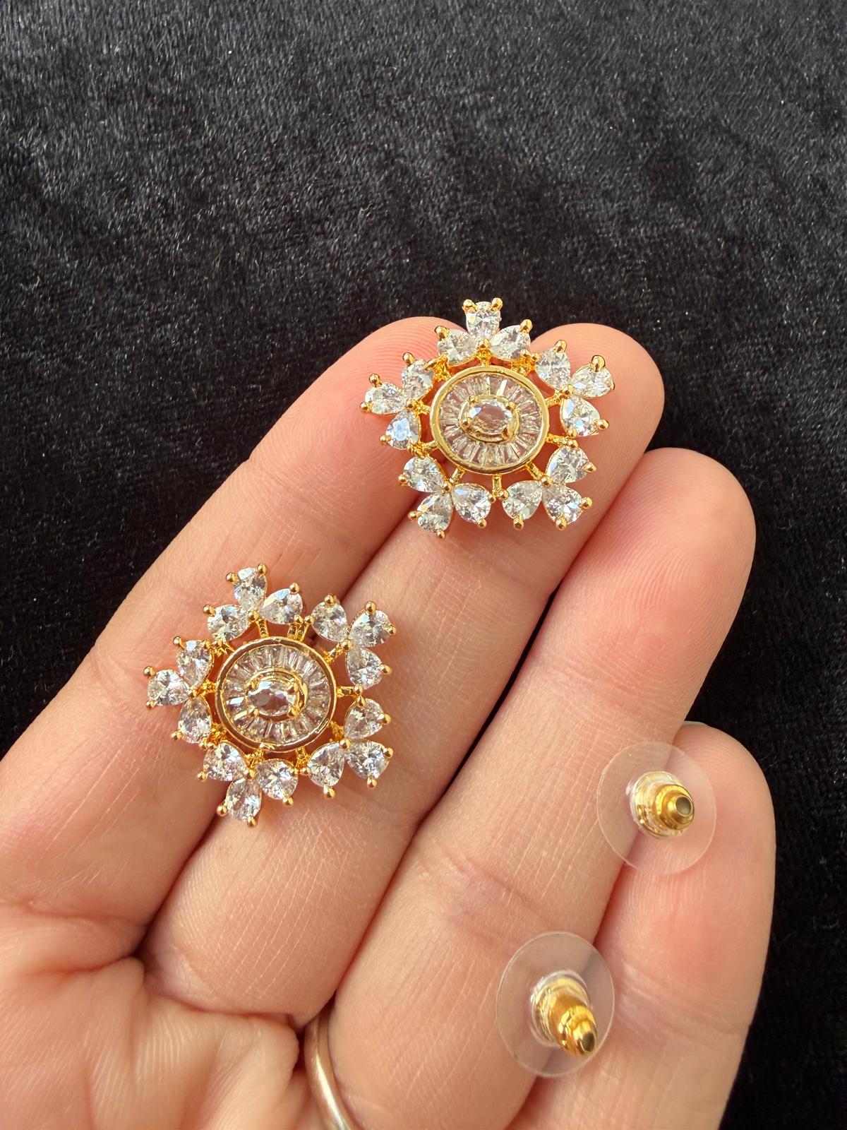 A close-up of a hand holding two gold-toned stud earrings with clear gemstone sunburst designs, with two additional earring backs visible on clear stands against a dark textured background.
