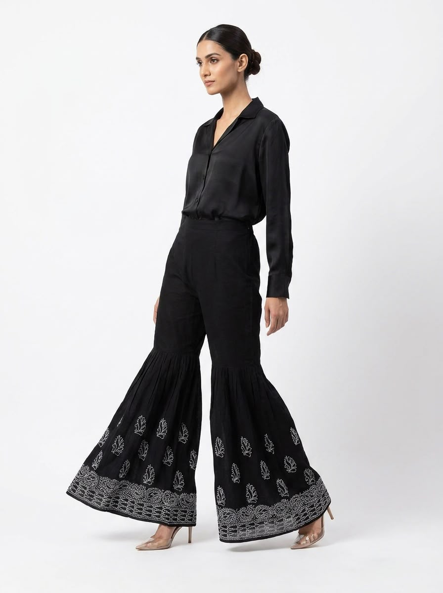 Side profile of a model showcasing the dramatic wide flare of jet black Chikankari embroidered sharara pants paired with a black shirt and heels against a white backdrop.