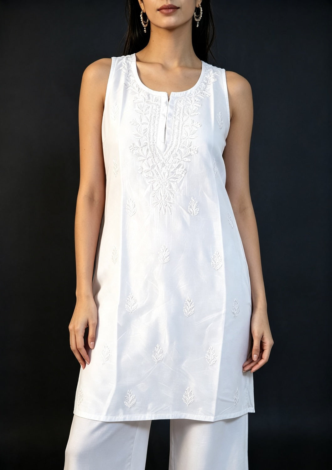 Medium close-up of a model wearing the white handcrafted sleeveless Chanderi silk Chikankari co-ord set, showcasing the fine embroidery detail against a solid dark backdrop.