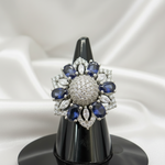 A floral-shaped silver ring brooch with deep blue oval-cut stones and clear pavé center and accents, displayed on a black cone stand against a gray gradient background. A white '2 cm' width measurement overlay is shown above the piece.