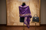 Purple shawl with floral patterns draped over a mannequin next to a black handbag on a small table against a marble wall.