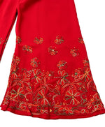 A close-up, flat-lay view of a red palazzo pant leg, featuring intricate, multi-color floral and vine embroidery at the hem and scattered motifs above. Against a plain white background.