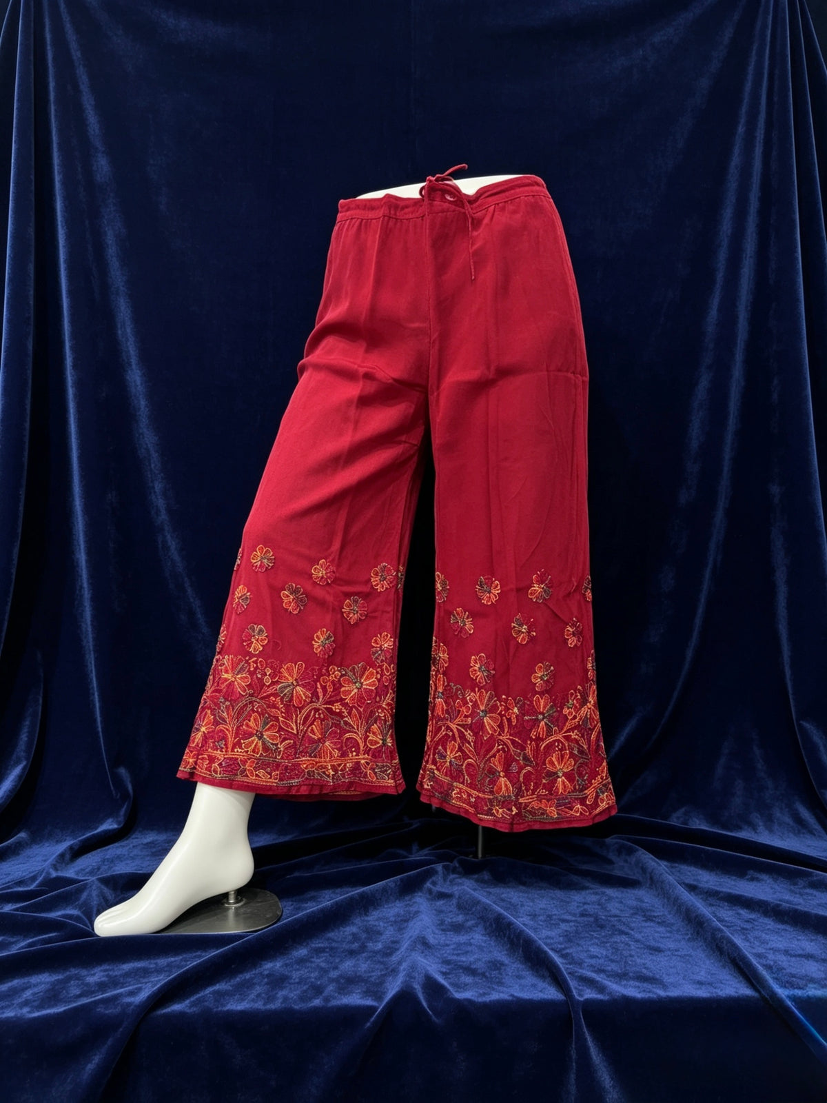 Red wide-leg cropped pants with ornate floral embroidery near the hem, displayed on a white mannequin leg against a dark blue velvet backdrop.