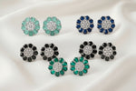 Six pairs of round flower-shaped stud earrings arranged in two rows on a draped white satin fabric. The top row features mint green, dark blue, and dark gray stones, while the bottom row features black, red, and pink stones, all set in silver-toned metal with clear central stones.