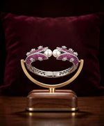 Luxury silver cuff bangle with deep purple stones and white pearls displayed on elegant gold and wood stand — Famaza Couture statement jewelry, silver-tone open cuff design with sparkling crystals and lustrous pearls, premium product photography with burgundy velvet background