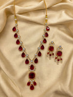 Ruby red pear & cushion-cut gemstone necklace with matching statement earrings, gold-tone finish with sparkling clear crystal accents — South Asian bridal jewelry laid on cream silk fabric.