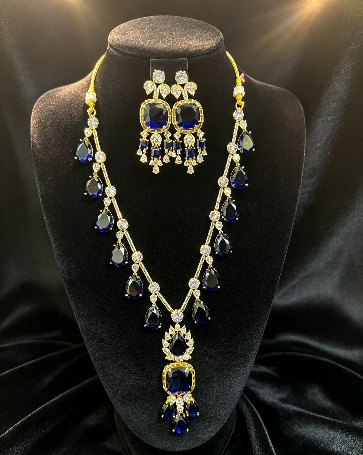 Sapphire blue pear & cushion-cut gemstone necklace with matching statement earrings, gold-tone finish with clear crystal accents — South Asian bridal & formal jewelry set on black display bust.