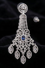 A close-up view of the back of an elegant chandelier earring featuring a central blue sapphire stone, intricate filigree metalwork, and sparkling diamond accents, shown with a clear rubber earring back.