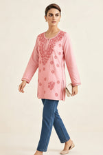 Full-length side view of a model walking while wearing a blush pink cotton short kurti featuring intricate pink embroidery, a keyhole neckline, and a square hem line, paired with matching trousers and holding a clutch.