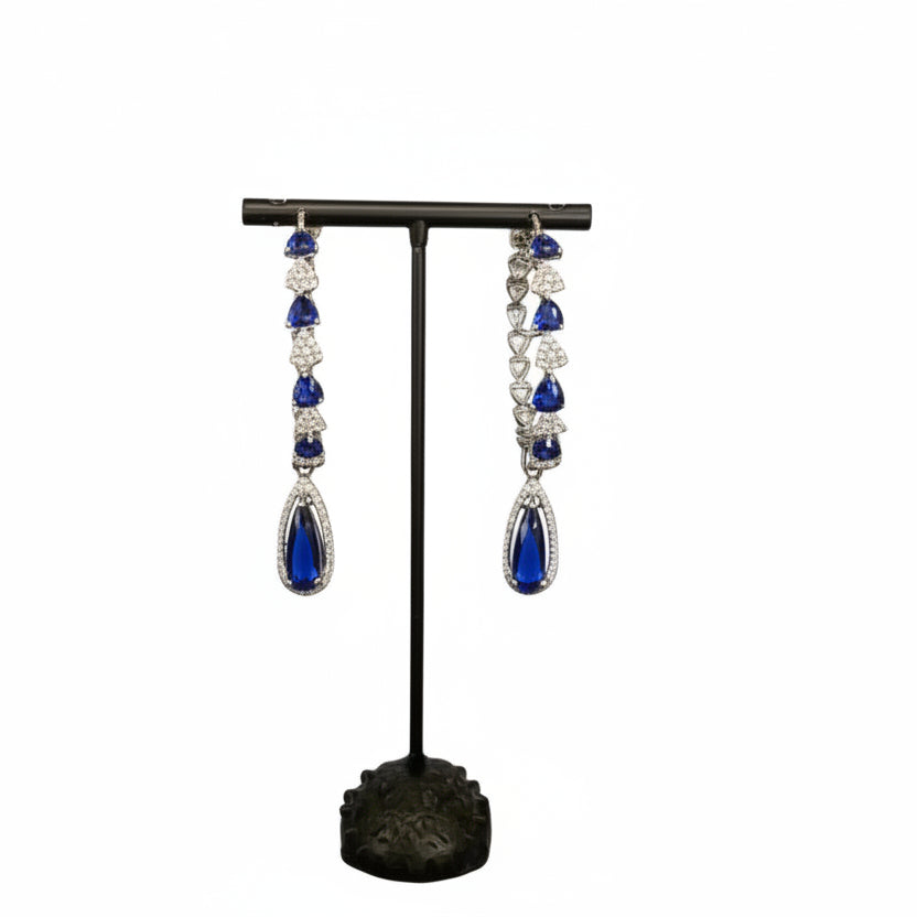 A pair of silver finish American diamond Jhumka bali earrings with green and clear stones on a stand.