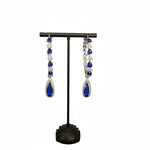 A pair of silver finish American diamond Jhumka bali earrings with green and clear stones on a stand.
