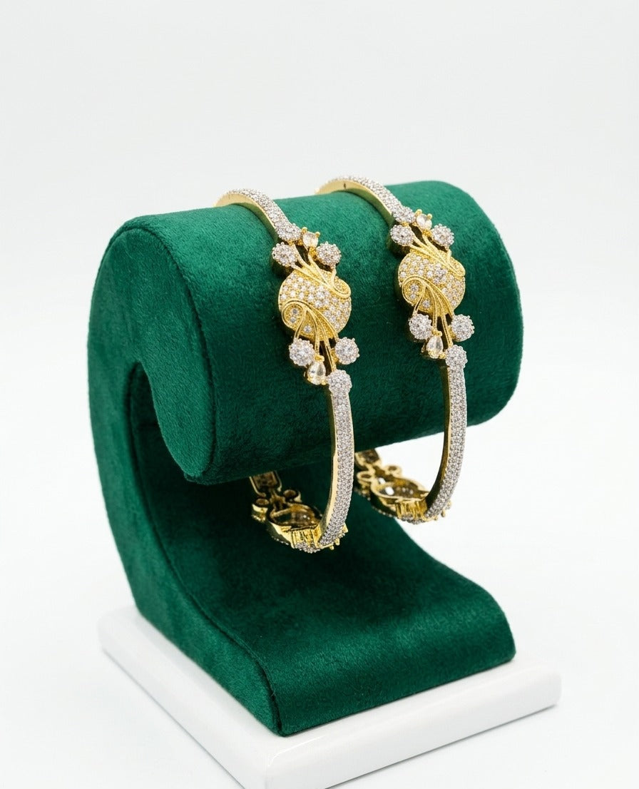 Pair of gold-plated American Diamond bangles featuring an intricate peacock floral motif and a pave-set stone band, displayed on a green velvet jewelry stand.