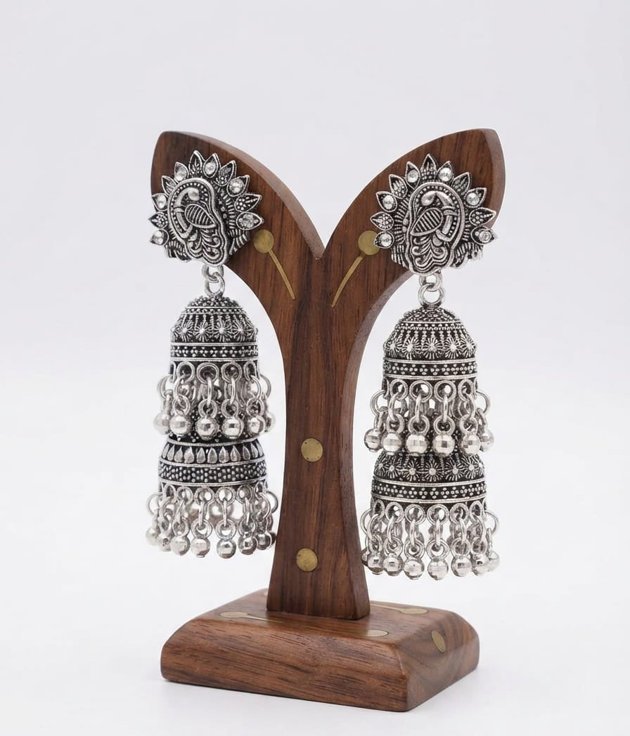 Pair of premium oxidized silver jhumka earrings featuring detailed peacock motifs on the top studs, multi‑layered bell design with dangling bead accents, displayed on a polished wooden earring stand against a plain white background.