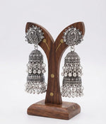 Pair of premium oxidized silver jhumka earrings featuring detailed peacock motifs on the top studs, multi‑layered bell design with dangling bead accents, displayed on a polished wooden earring stand against a plain white background.