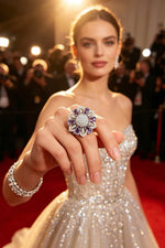 Silver-tone floral halo ring with deep purple stones and sparkling AD center, held forward on hand against glamorous red carpet backdrop.