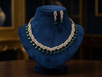 A gold necklace and matching drop earrings with green teardrop gemstones, displayed on a dark blue velvet jewelry bust against a dark, elegant background.