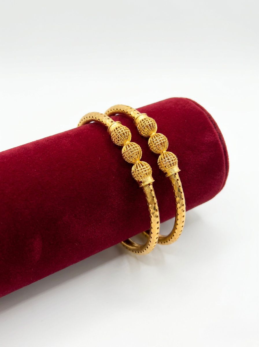 A pair of micro gold-plated copper bangles featuring three hollow filigree orbs and a textured band, displayed on a red velvet jewelry stand against a white background