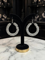 Pair of silver-tone crystal-encrusted teardrop hoop earrings on black velvet stand with gold base — Famaza Couture bridal luxury jewelry.